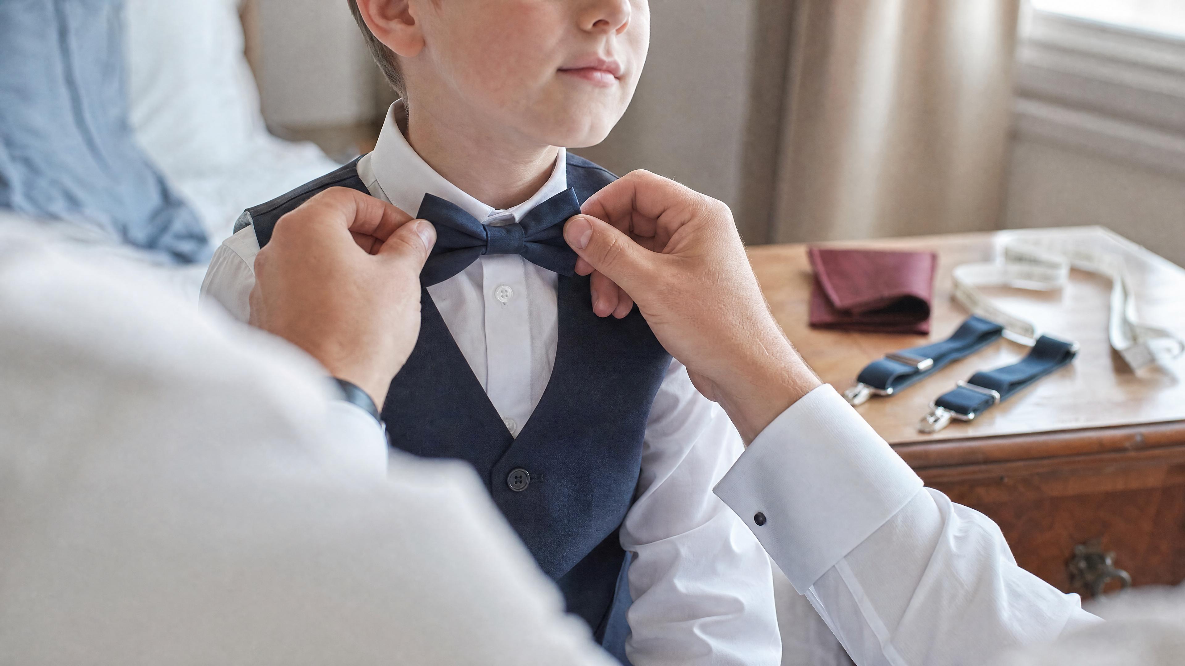 Un adulte ajuste un nœud papillon sur un enfant en chemise blanche, avec un mètre ruban visible sur une commode, dans une chambre lumineuse avant une cérémonie Immagine